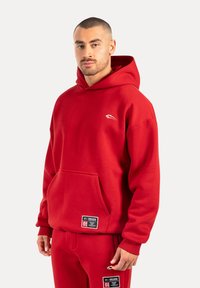 Sudadera con capucha roja y gran bolsillo frontal, fabricada en material suave. Presenta un pequeño logo blanco en el lado izquierdo del pecho y pantalones a juego.