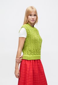 Rokām adīts bezr piedurknēm džemperis spilgti zaļā krāsā ar pītas adījuma rakstu un ribotu apakšmalu, saskaņots ar baltu kreklu un sarkanu svītrainu svārku.