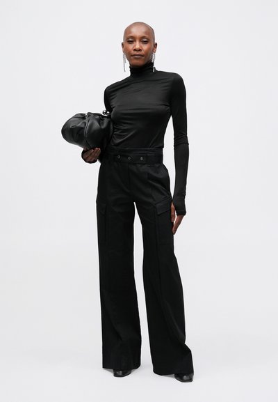 Cămașă neagră cu mâneci lungi, ajustată, și pantaloni largi cu talie înaltă, amândouă dintr-o țesătură netedă. Modelul ține un plic negru, cu un design moale și rotunjit.