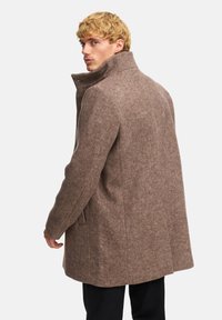 Abrigo de mezcla de lana marrón con un cuello alto, acabado texturizado y bolsillos laterales, llevado por un modelo de espaldas contra un fondo blanco.