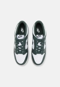 Des baskets Nike blanches et vertes présentent une tige en cuir lisse, un orteil perforé et des lacets plats. Le logo de la marque est visible sur la languette et sur le côté.