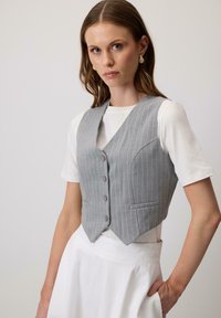 Pilkas dryžuotas liemenės modelis, su laisvai prilipusiu dizainu, turintis gilią V formos iškirptę, penkis mygtukus ir dvi priekinės kišenes, uždėtas ant baltos marškinių.