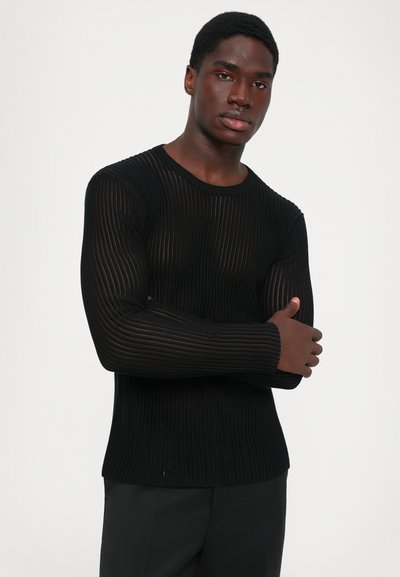 Νεαρός άνδρας με σταυρωμένα χέρια, φορώντας μαύρο ριγέ ημιδιαφανές πουλόβερ με μακρύ μανίκι και μαύρο παντελόνι, μπροστά σε λευκό φόντο.