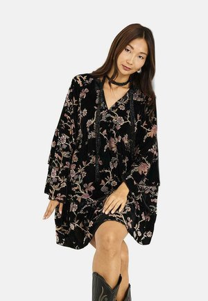 Joven mujer con un vestido de terciopelo negro con flores, de mangas largas y un collar, posando con una rodilla doblada y botas de vaquero negras.