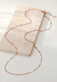 Lange, roségoldene Kettenhalskette mit strukturierten Gliedern, auf einer hellen Steinoberfläche drapiert, die ein längliches, fließendes Design präsentiert.