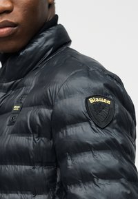 Κοντινό πλάνο ατόμου που φοράει μαύρο μπουφάν τύπου puffer με το λογότυπο Blauer στο μανίκι, πάνω σε απλό ανοιχτό φόντο.