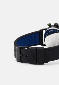 Correa de reloj de silicona negra con acentos azules y perforaciones. Esfera redonda con respaldo de acero inoxidable y detalle en la corona de color verde. Logotipo de Adidas visible.