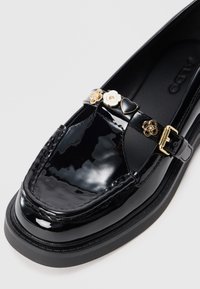Black patent leather shoe with a rounded toe, featuring a decorative strap adorned with gold hardware and floral accents. Thick rubber sole.