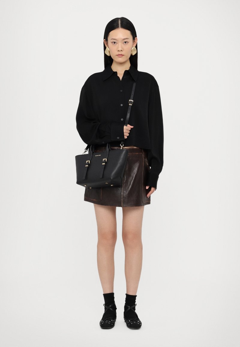 Borsa a tracolla in pelle nera con dettagli dorati, tracolla regolabile e design strutturato, abbinata a una camicia nera oversize e una gonna in pelle marrone.