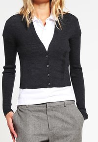 Svart ribbad cardigan med långa ärmar, V-ringning och sex knappar. Buren över en vit skjorta med krage, i kombination med grå byxor.
