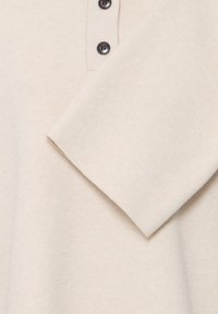 Pull en maille beige à manches longues, trois boutons noirs sur le haut du torse, texture douce, montré en gros plan.