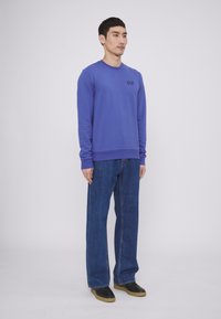 Felpa viola a maniche lunghe con logo sul petto, abbinata a jeans in denim blu a taglio loose e scarpe nere con suola in gomma.