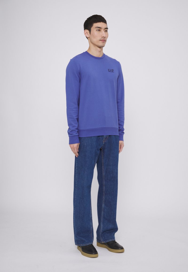 Felpa viola a maniche lunghe con logo sul petto, abbinata a jeans in denim blu a taglio loose e scarpe nere con suola in gomma.