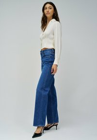 Blusa blanca corta con cuello, combinada con jeans de mezclilla azul de pierna ancha y cintura alta, y tacones negros de punta. Fondo neutro.