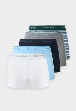 Młody mężczyzna stoi boso, ubrany w czarne bokserki Calvin Klein na jednolitym jasnym tle.