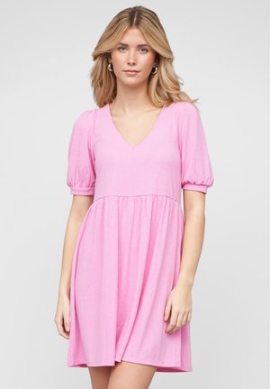 Ung kvinne med blondt hår ikledd en rosa kortermet V-halskjole, stående mot en enkel lys grå bakgrunn.