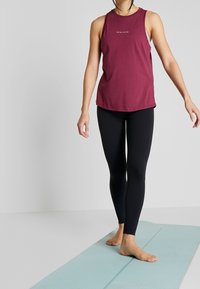 Top senza maniche color marrone amarena con una semplice stampa frontale, abbinato a leggings neri. L'outfit è completato da piedi nudi su un tappeto di un blu chiaro.