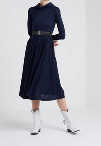 Knälång marinblå klänning med draperad halsringning, långa ärmar och figursydd midja, tillsammans med vita cowboystövlar med sidotasslar.