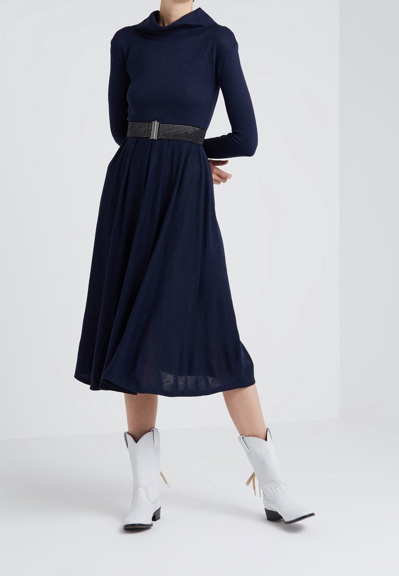 Knälång marinblå klänning med draperad halsringning, långa ärmar och figursydd midja, tillsammans med vita cowboystövlar med sidotasslar.