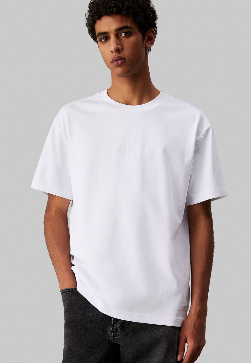 Camiseta de algodón blanca con mangas cortas y cuello redondo. Sutil logotipo "CK" en relieve en el pecho. Combinada con jeans oscuros.