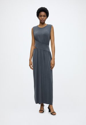 Woman with short curly hair wearing a sleeveless dark gray maxi dress with side cutout, standing and wearing black heeled sandals.