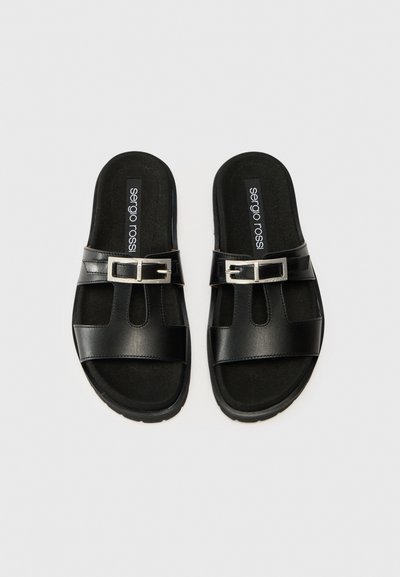 Une paire de sandales en cuir noir à glissière avec des boucles en argent et des semelles rembourrées sur un fond uni.