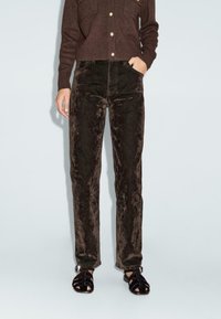 Pantaloni larghi in velluto marrone con vita alta. Tessuto testurizzato con una leggera lucentezza, abbinati a sandali neri con lacci.