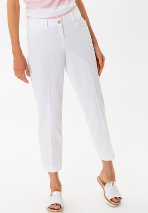 Femme portant un pantalon blanc sur mesure longueur cheville avec un bouton et des poches, associé à des sandales argentées et blanches à bout ouvert.