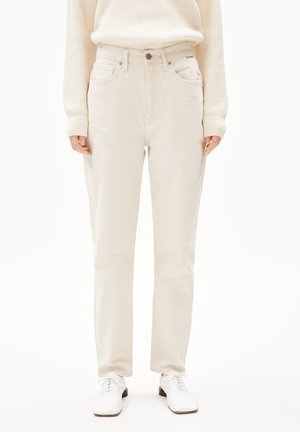 Osoba ubrana w kremowe dżinsy z wysokim stanem, kremowy prążkowany sweter oraz białe sznurowane buty na gładkim białym tle.