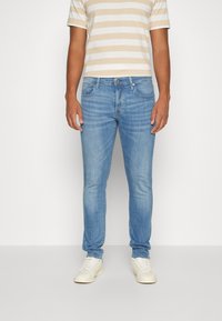 Man som bär ljusblå slim-fit jeans, beige och vit randig kortärmad skjorta samt vita sneakers, stående mot en enfärgad bakgrund.