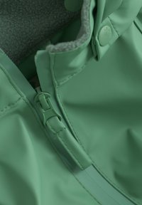 Groene waterdichte jackdetail met een gladde buitenlaag, een donzige grijze voering en een bijpassende plastic rits met drukknoopsluiting.