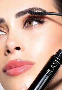 Mascara nera in un tubetto tenuto da una persona mentre lo applica sulle ciglia. Pelle liscia, labbra lucide e sopracciglia definite con un volume evidente.