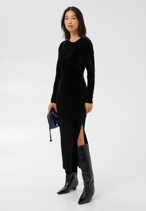 Femme portant une longue robe en velours noire avec une fente sur le côté, des bottes noires montant jusqu'au genou, tenant un petit sac à main bleu marine, debout sur un fond uni.