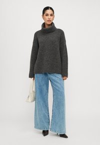 Maglione grigio scuro a maglia con colletto alto, abbinato a jeans a gamba larga azzurri. La modella tiene una piccola borsa bianca e indossa scarpe nere a punta.