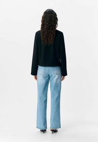 Donna con lunghi capelli ricci che indossa un blazer nero, jeans dritti azzurri chiari e sandali con tacco neri, in piedi di spalle su uno sfondo bianco.