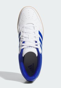 Zapatilla deportiva blanca con detalles en azul, que presenta una puntera perforada, tres franjas azules y una suela de goma texturizada.