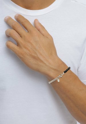 Bracelet en perles noires avec détails en chaîne argentée et fermoir, porté sur une main contre un fond de chemise blanche.