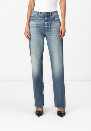 Woman wearing high-waisted, straight-leg faded blue jeans with front button closure, paired with black pointed heels with buckles.