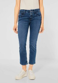 Jean en denim bleu avec une coupe slim, taille mi-haute et deux poches avant. Les jambes sont légèrement raccourcies avec des ourlets bruts. Associé avec des baskets blanches.