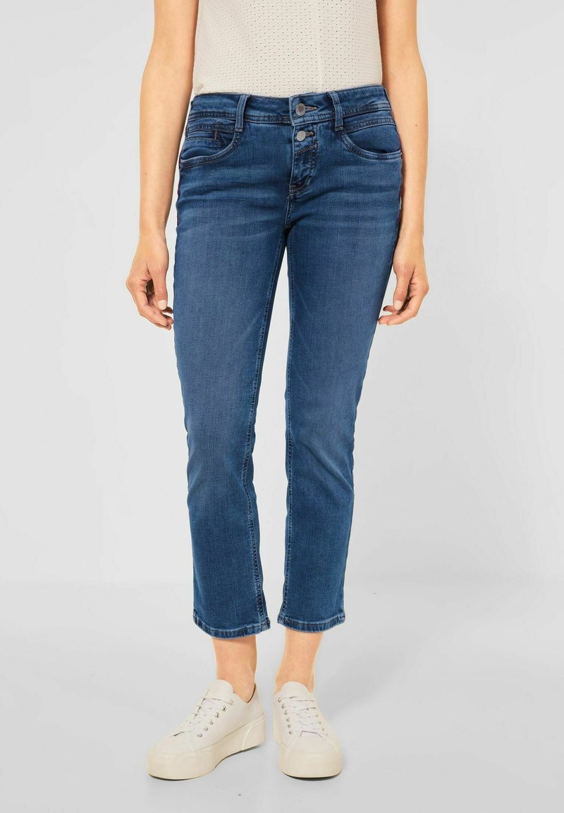 Jean en denim bleu avec une coupe slim, taille mi-haute et deux poches avant. Les jambes sont légèrement raccourcies avec des ourlets bruts. Associé avec des baskets blanches.