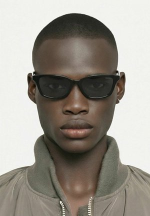 Gros plan d'une personne à la peau foncée portant des lunettes de soleil rectangulaires noires et une veste grise à col montant sur un fond clair.