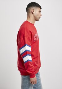 Rotes Sweatshirt mit weißen und blauen Streifenakzenten an den Ärmeln, mit einer strukturierten Oberfläche und gerippten Bündchen, kombiniert mit lässiger Jeans.