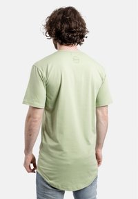 Camisa de manga corta verde claro con dobladillo redondeado y detalle de logo en la parte trasera. La tela parece suave y lisa, enfatizando un corte relajado.