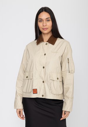 Kobieta o długich ciemnych włosach, ubrana w beżową zapinaną na guziki kurtkę z brązowym kołnierzem i dużymi przednimi kieszeniami, stojąca na jednolitym tle.