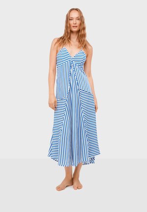 Mujer de pie descalza, vistiendo un vestido maxi sin mangas a rayas azules y blancas, con escote en v y falda fluida, sobre un fondo liso.