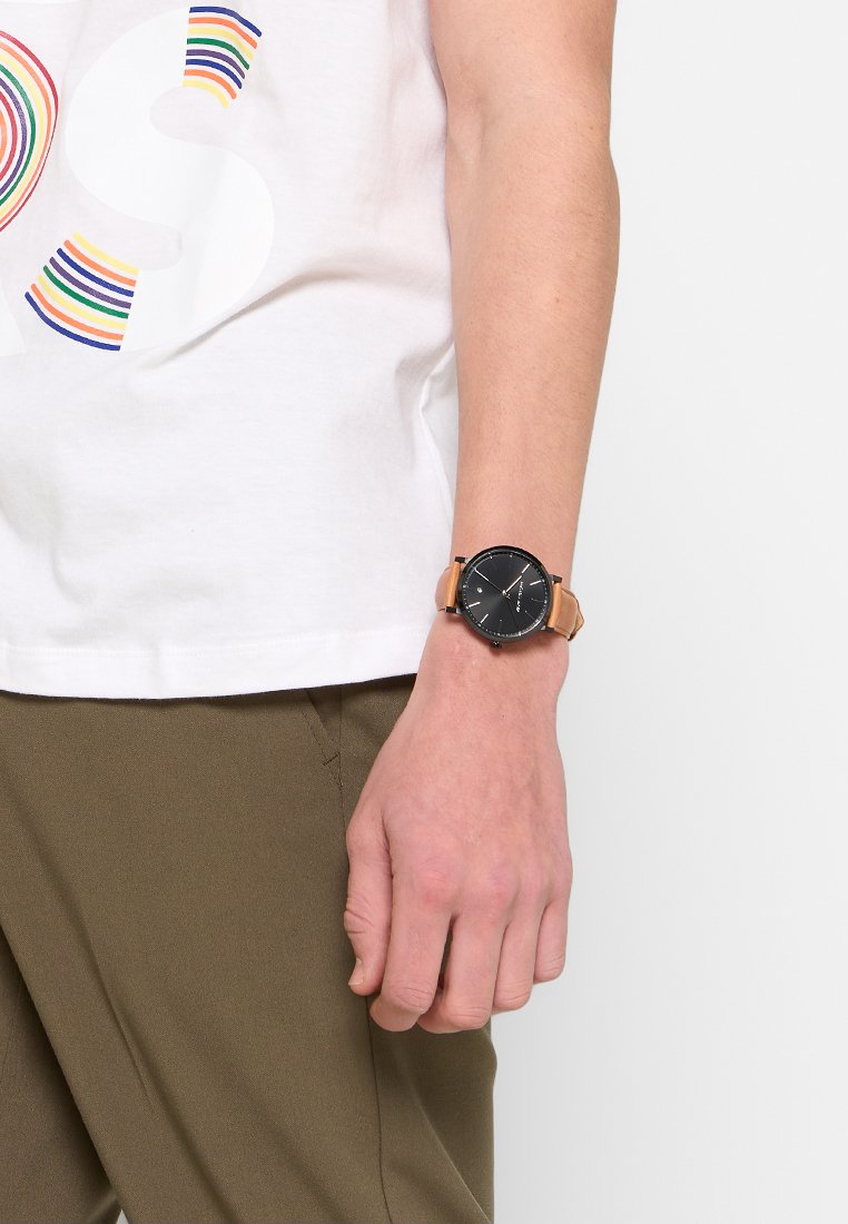 Zwart wijzerplaat horloge met een ronde oppervlakte, goudkleurige accenten en een tan lederen band. Draagt om een pols met een olijfgroen broek en een wit shirt.