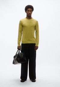 Φούτερ σε λάιμ πράσινο με στρογγυλή λαιμόκοψη, φορεμένο με μαύρο παντελόνι φαρδιάς γραμμής. Μαύρη υφασμάτινη τσάντα με διπλούς χειρολαβές, τοποθετημένη σε λευκό φόντο.