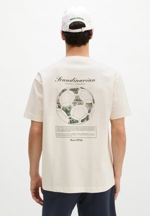 Άνδρας που φορά λευκό καπέλο και κρεμ μπλουζάκι με μια παλιά μπάλα ποδοσφαίρου και το κείμενο «Scandinavian Football Community» στην πλάτη.