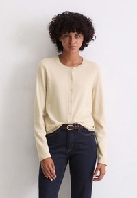 Helles beiges Cardigan mit rundem Ausschnitt, Knopfschluss und langen Ärmeln. Kombiniert mit dunklen Jeans und einem braunen Gürtel. Glatte Textur.