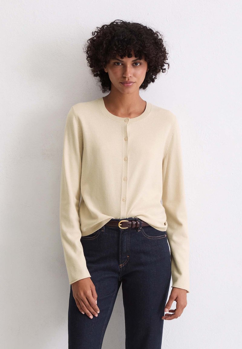 Helles beiges Cardigan mit rundem Ausschnitt, Knopfschluss und langen Ärmeln. Kombiniert mit dunklen Jeans und einem braunen Gürtel. Glatte Textur.